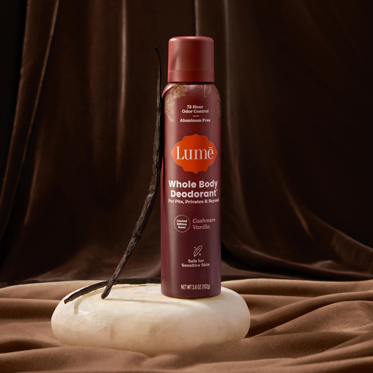 A can of Lume spray deodorant with vanilla beans leaning against it, in front of luxe fabric.